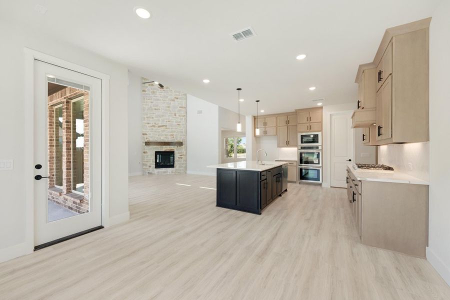Open-concept kitchen with dark island, tan cabinets, stainless appliances, and stone fireplace in adjoining living area