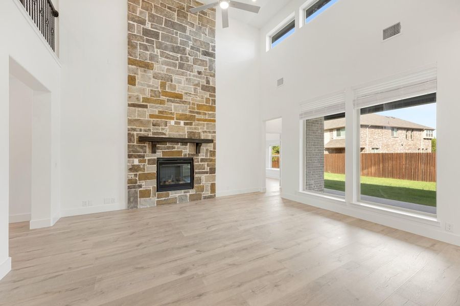 Open living room with two-story stone fireplace, light wood floors, and large windows overlooking backyard