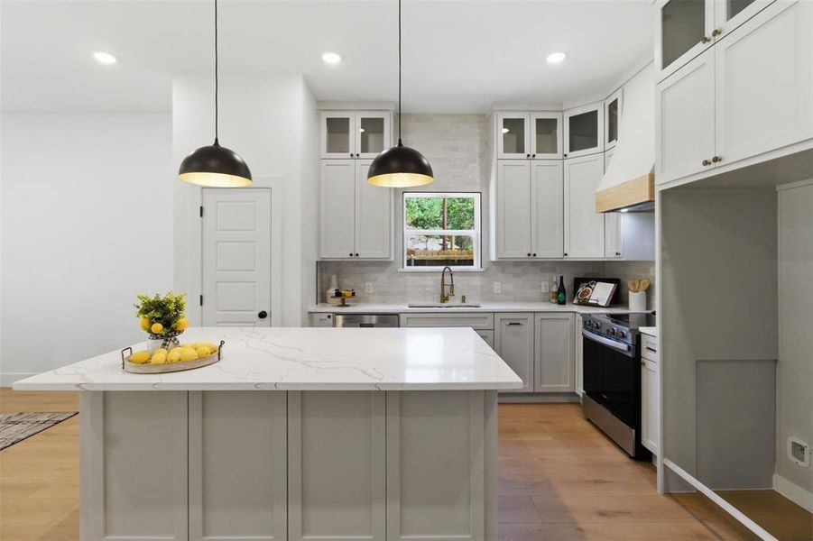 Kitchen featuring a substantial island with a light-toned countertop, white cabinetry with glass upper doors, wood-finish flooring, a stainless steel range, and two pendant light fixtures