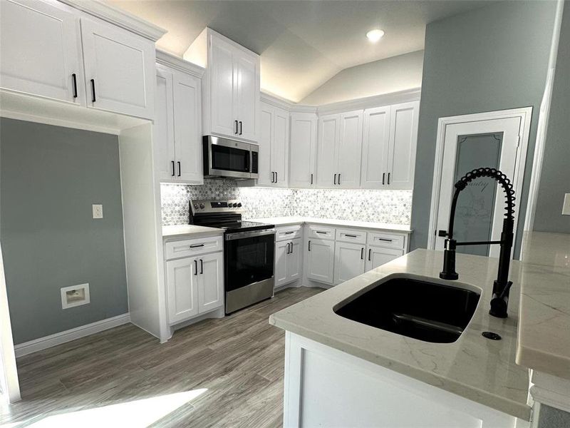 Kitchen with light stone countertops, backsplash, stainless steel appliances, vaulted ceiling, and white cabinets