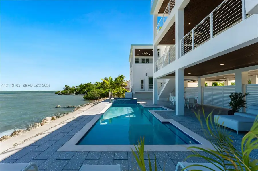 Waterfront exterior featuring a multi-level structure with recessed balconies, horizontal cable railings, a rectangular swimming pool with an integrated spa, and expansive stone-tiled patio decking Waterfront exterior featuring a multi-level structure with recessed balconies, horizontal cable railings, a rectangular swimming pool with an integrated spa, and expansive stone-tiled patio decking