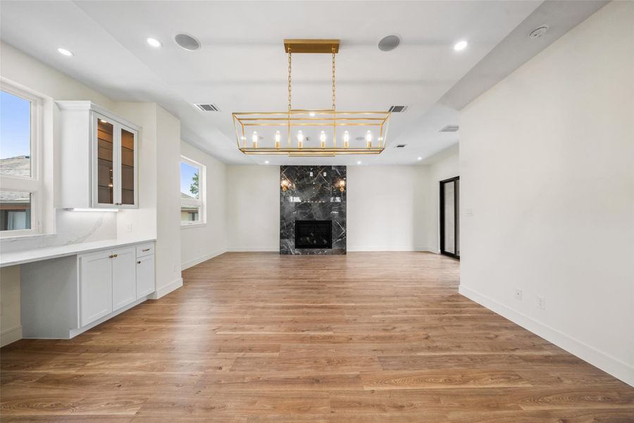 Open-concept floor plan features a sleek wood floor, a modern chandelier, and a striking black marble fireplace. Natural light streams in through large windows, complementing the elegant white cabinetry and ample counter space, perfect for entertaining.