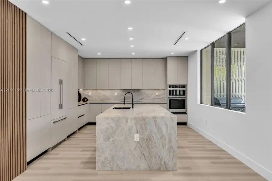 Kitchen featuring floor-to-ceiling cabinetry, a waterfall stone island, integrated stainless steel appliances, wood-finish flooring, and slatted wood accent detailing Kitchen featuring floor-to-ceiling cabinetry, a waterfall stone island, integrated stainless steel appliances, wood-finish flooring, and slatted wood accent detailing