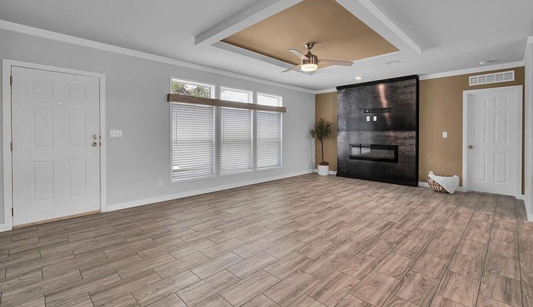 Unfurnished living room featuring ornamental molding, ceiling fan, light wood-style floors, a large fireplace, and a raised ceiling Unfurnished living room featuring ornamental molding, ceiling fan, light wood-style floors, a large fireplace, and a raised ceiling