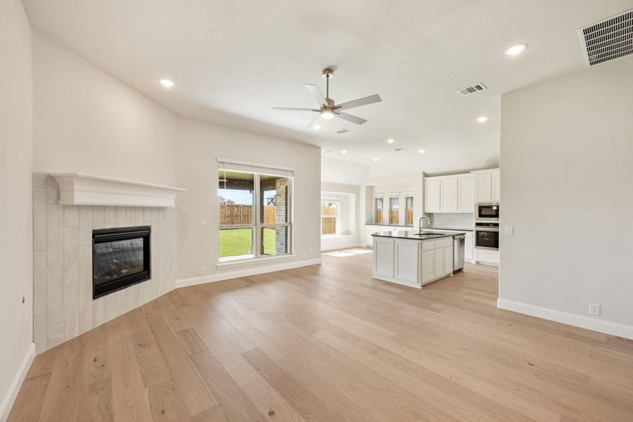 Open living area with tile fireplace, ceiling fan, and hardwood floors flowing into white kitchen with island Open living area with tile fireplace, ceiling fan, and hardwood floors flowing into white kitchen with island