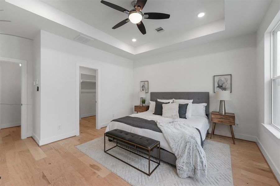 Spacious and well-lit primary bedroom features custom vaulted modern tray ceilings, modern ceiling fan, recessed lighting, engineered hardwood floors, and a neutral color palette. There are two doorways, leading to your walk in closet and private en suite.