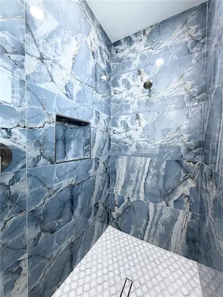 Shower stall featuring full-height blue-toned stone-patterned wall tiles, integrated recessed shelving, white geometric mosaic floor tiling, and a metallic shower fixture