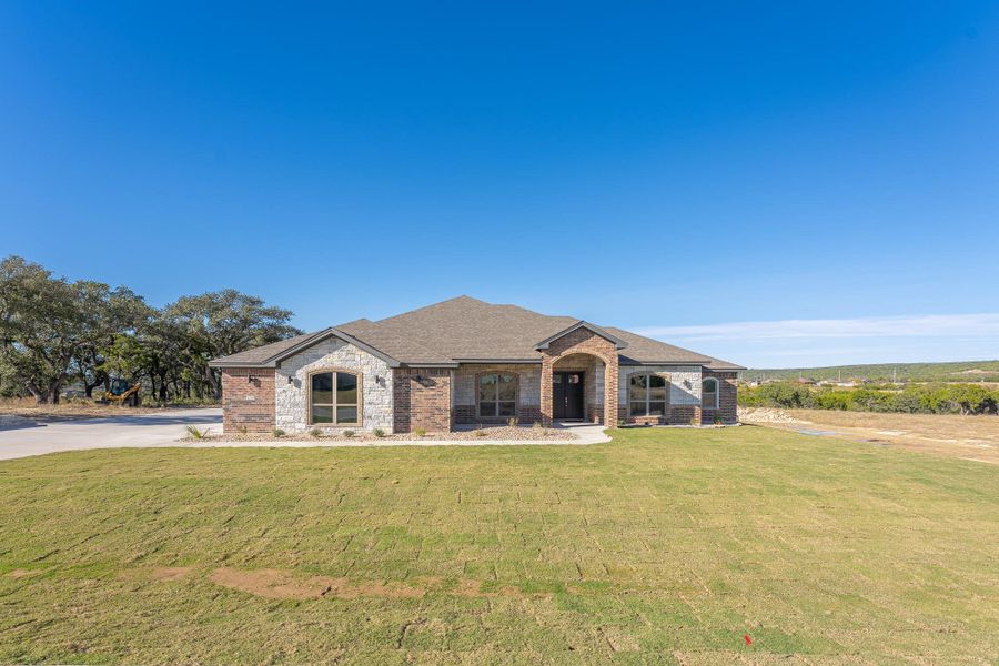 View of front facade featuring brick siding, a front yard, a shingled roof, and stone siding View of front facade featuring brick siding, a front yard, a shingled roof, and stone siding