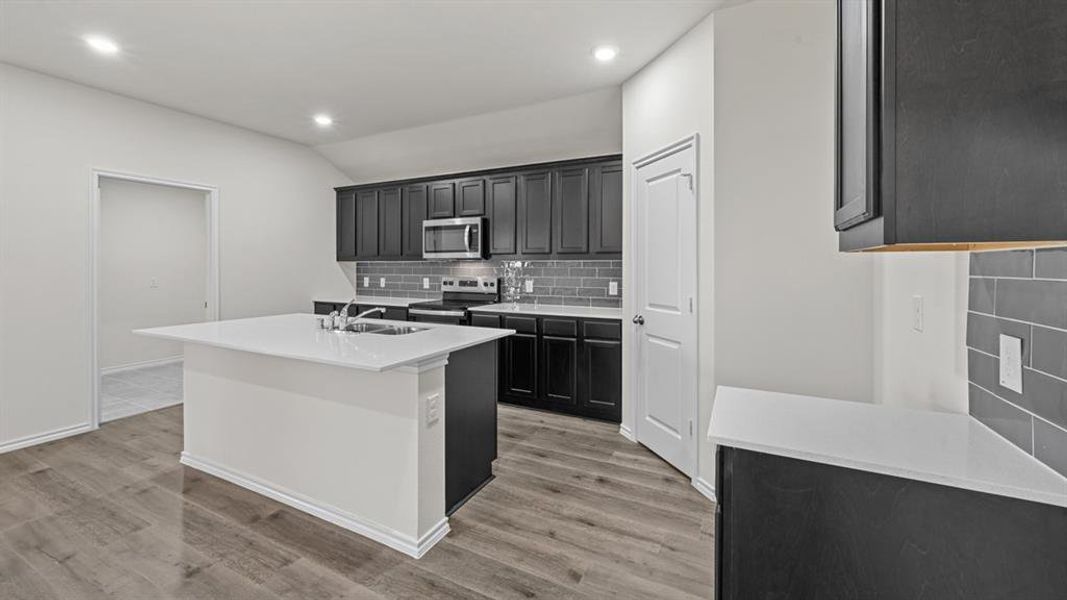 Kitchen featuring a central island with a sink, dark wood-finish cabinetry, light-toned countertops, stainless steel appliances, and a gray subway tile backsplash