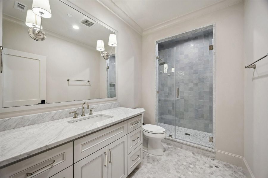 Beautifully appointed ensuite bath featuring Carrara mosaic tile flooring, marble slab counter, custom cabinetry, and a frameless glass-enclosed shower with striking contrasting tile to the ceiling for added visual interest. Clean, classic finishes create a bright and inviting space.