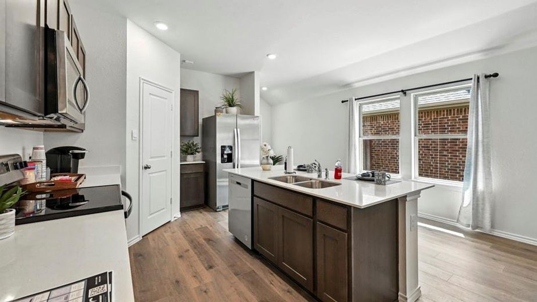 Open-concept kitchen featuring an island with a dual-basin sink, stainless steel appliances, wood-finish flooring, and ample natural light from multiple windows
