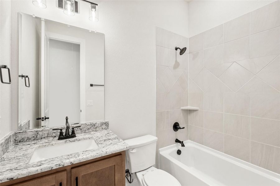 Bathroom with granite vanity, wood cabinets, and tiled tub-shower combo with dark fixtures