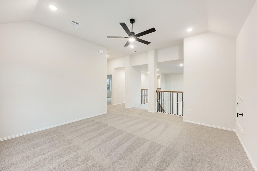 Open upstairs loft with beige carpet, black ceiling fan, and iron stair railing along open landing Open upstairs loft with beige carpet, black ceiling fan, and iron stair railing along open landing