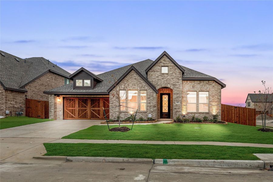 Single-story brick home with arched entry, wood garage doors, and manicured lawn at dusk