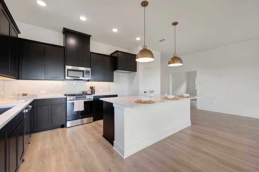 Kitchen featuring a kitchen island, stainless steel appliances, pendant lighting, dark cabinetry, and light wood-type flooring Kitchen featuring a kitchen island, stainless steel appliances, pendant lighting, dark cabinetry, and light wood-type flooring