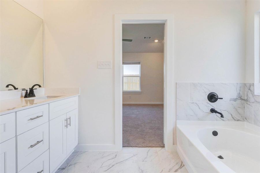 Relax and unwind in the private primary bath showcasing a soaking tub with marble-style tile surround and contemporary black fixtures. The layout offers a seamless connection to the primary bedroom, enhancing flow and functionality. Neutral finishes, soft natural light, and clean lines create a calm, inviting retreat. This well-appointed space balances luxury and practicality, perfect for starting and ending your day in comfort.
