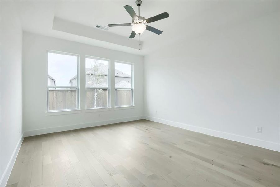 Spacious room featuring three large windows, light wood-finish flooring, a contemporary ceiling fan with integrated lighting, a tray ceiling detail, and crisp white walls with matching baseboards