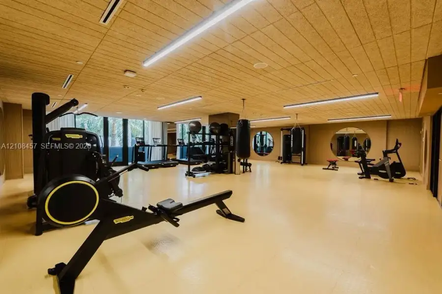 Spacious fitness room featuring wood-slat ceiling, recessed linear lighting, circular wall mirrors, and textured resilient flooring Spacious fitness room featuring wood-slat ceiling, recessed linear lighting, circular wall mirrors, and textured resilient flooring