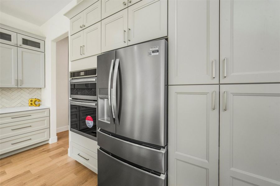 This kitchen features modern white cabinetry, a sleek stainless steel refrigerator, and double wall ovens. The light wood flooring and herringbone backsplash add warmth and style to the space.