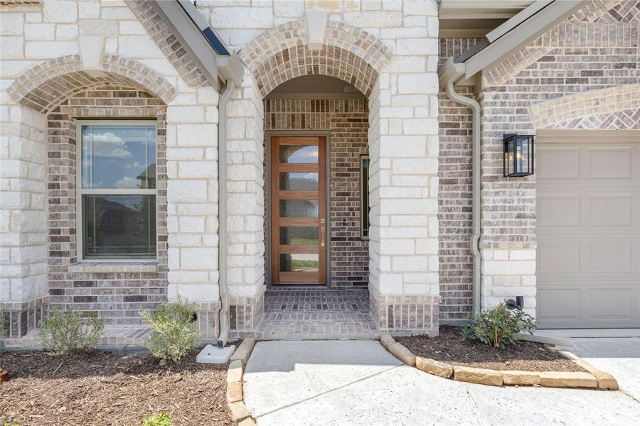 Home entrance with arched brick and stone surround, wood front door with glass panels, and wall lantern nearby.