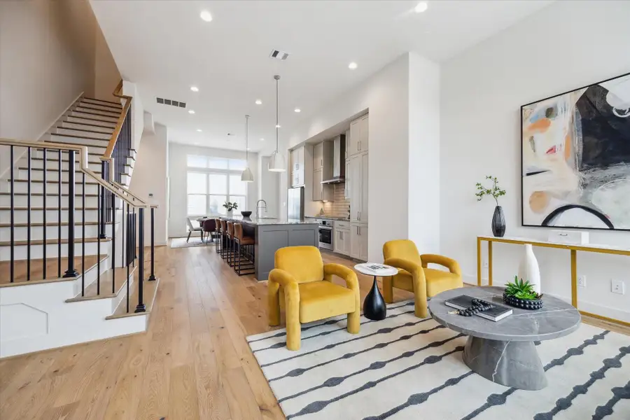 This modern living space features an open concept design with a sleek kitchen, island seating, and elegant pendant lighting. The living area includes vibrant yellow chairs, a stylish rug, and contemporary artwork. Natural wood flooring and a staircase with black railings add warmth and sophistication.