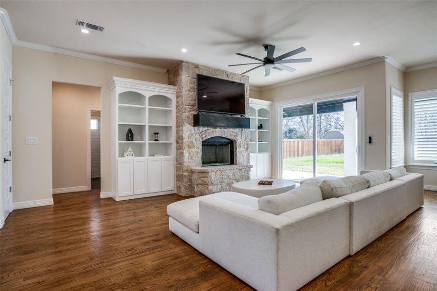 Living room with ceiling fan, a stone fireplace, crown molding, and dark wood-type flooring