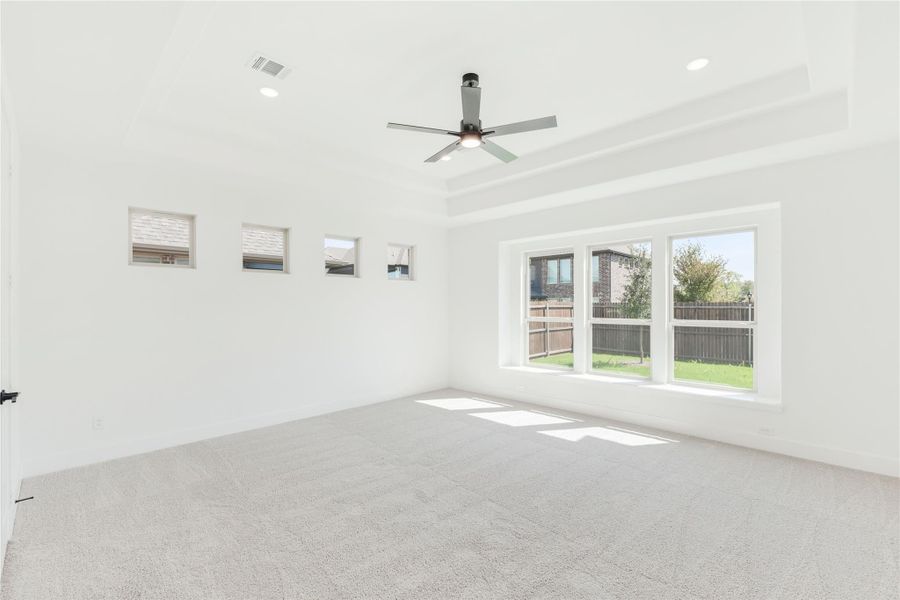 Empty bedroom with tray ceiling, ceiling fan, carpet flooring, and large windows overlooking backyard