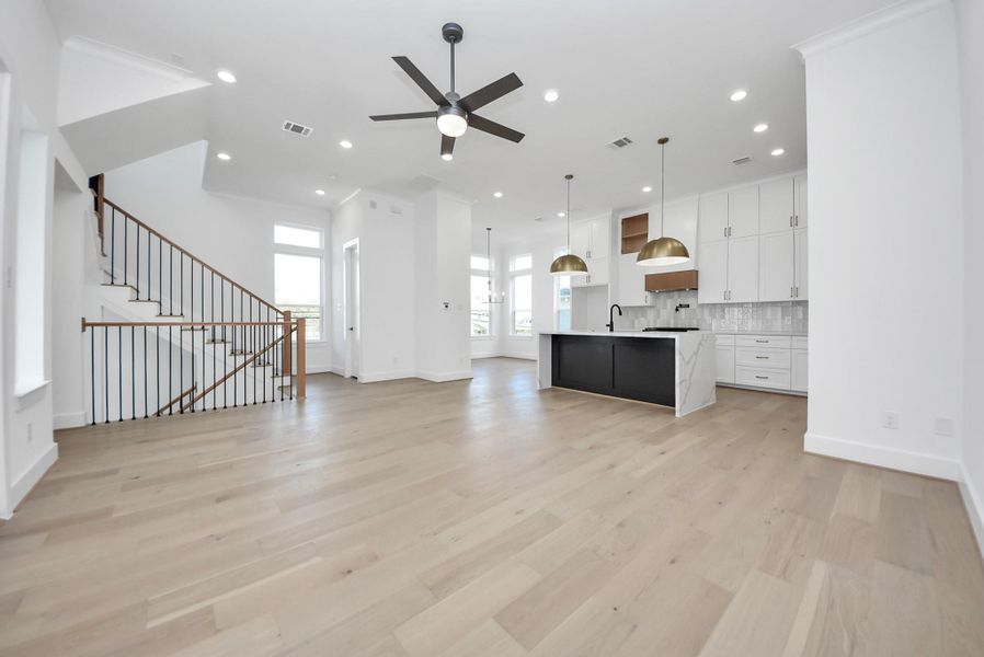 An open-concept living area showcasing light wood floors and a refined kitchen design. The large island features a marble countertop with a contrasting dark base, complemented by gold pendant lighting, white cabinetry, and a modern staircase—all illuminated by abundant natural light.