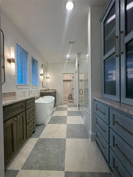 Spacious bathroom featuring checkerboard tile flooring, a freestanding soaking tub, dual vanity areas with stone countertops, a glass-enclosed shower, and built-in cabinetry with reeded glass inserts