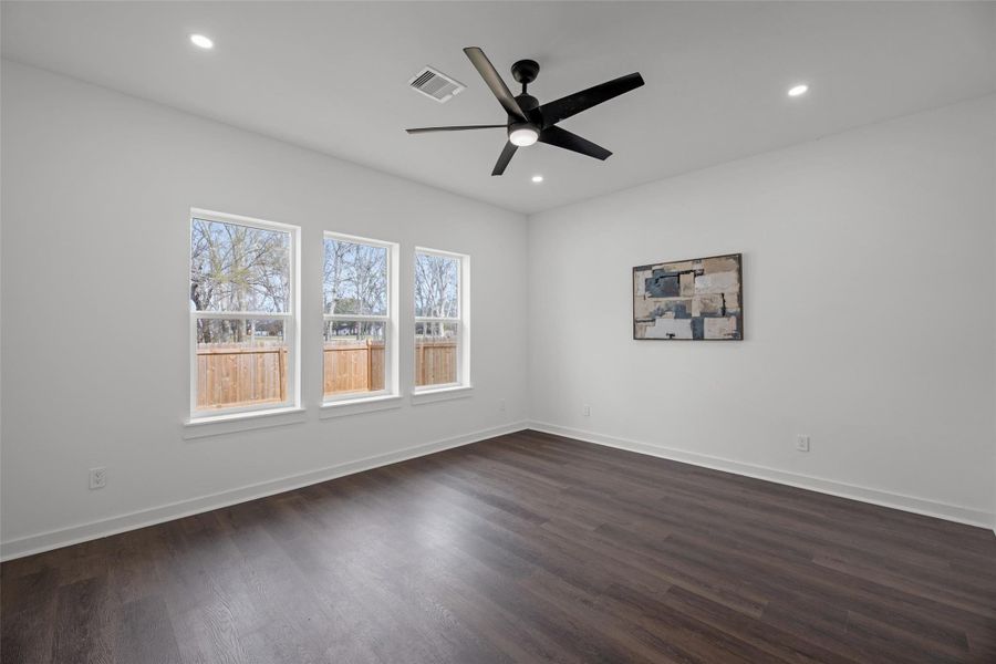 Lighted, modern room with dark wood flooring, white walls, and recessed lighting. Features a ceiling fan, three large windows for natural light, and a piece of wall art. Lighted, modern room with dark wood flooring, white walls, and recessed lighting. Features a ceiling fan, three large windows for natural light, and a piece of wall art.