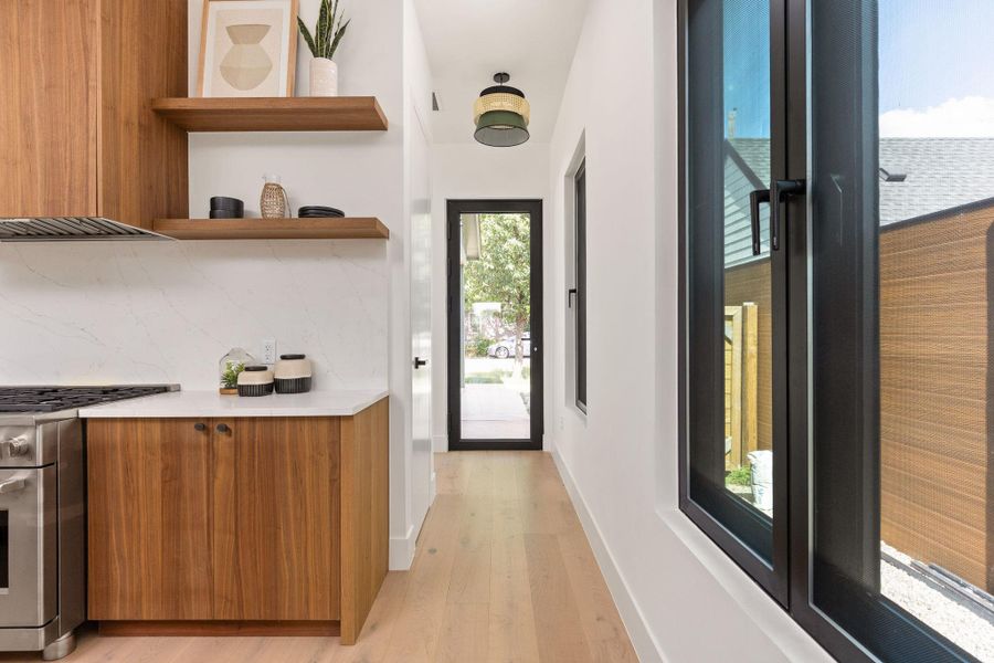 A well-appointed kitchen features rich wood cabinetry, floating shelves, and an adjoining brightly lit hallway with a glass-paneled entry door.