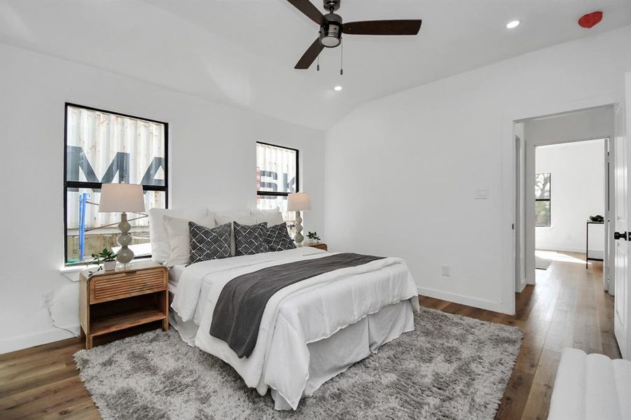 This is a modern and minimalist bedroom featuring clean, white walls with a single abstract artwork, a ceiling fan, recessed lighting, a plush area rug over hardwood floors, and a bed dressed in neutral linens. There's a doorway leading to another room.