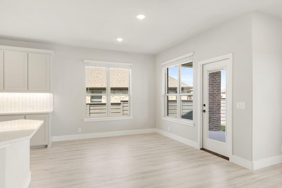Bright dining area with light wood floors, two windows, and a glass back door next to white kitchen cabinets