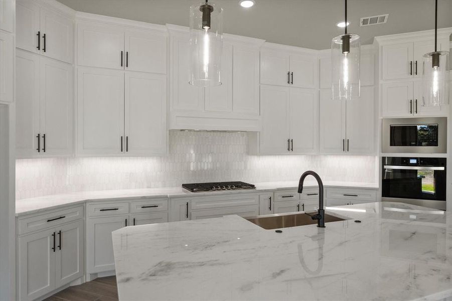 Kitchen featuring quartz countertops, marble tile backsplash, huge island with apron sink & storage cabinets under overhang, ss appliances, undercounter lighting, hidden outlets and pendant lighting.