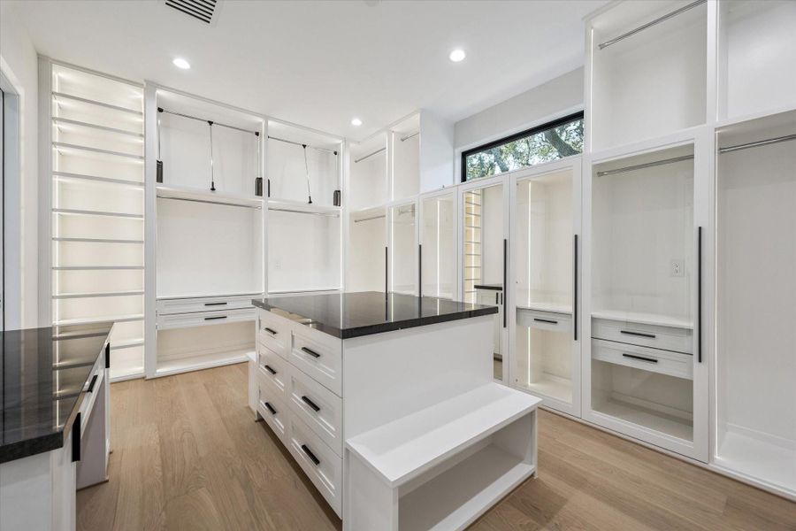 The expansive walk-in closet is outfitted with custom storage, including lighted glass cabinetry, abundant hanging space, and sleek drawers. A central island with quartz countertop adds storage and workspace, while full-length mirrors and warm wood flooring enhance the boutique-like experience.