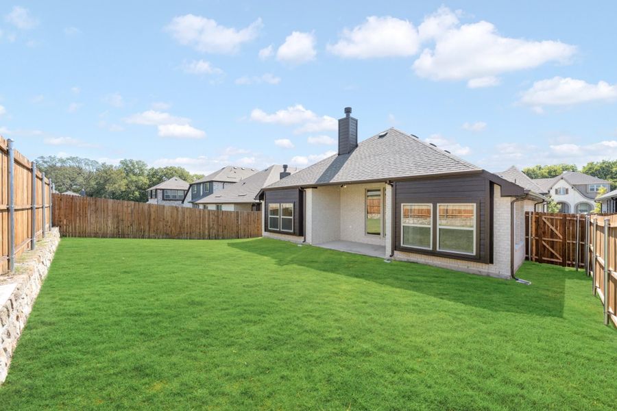 Backyard of single-story home with brick exterior, covered patio, large green lawn, and wood privacy fence.