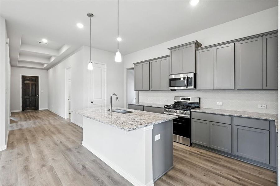 Kitchen with gray cabinets, stainless steel appliances, light stone countertops, a center island with sink, and light wood-style flooring