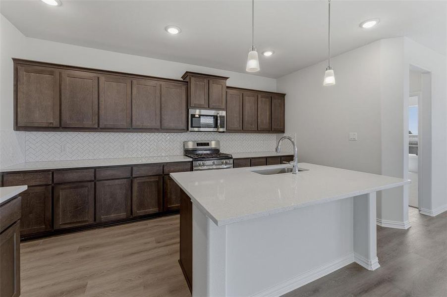 Modern kitchen featuring a central island with an undermount sink, dark wood cabinetry, white herringbone tile backsplash, stainless steel appliances, and wood-finish flooring