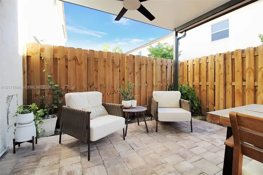 Covered outdoor patio featuring stone-paver flooring, a ceiling-mounted fan, and natural wood privacy fencing Covered outdoor patio featuring stone-paver flooring, a ceiling-mounted fan, and natural wood privacy fencing