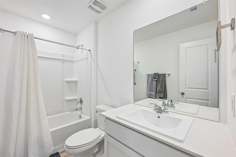 Full bathroom featuring a white tub/shower combination with a built-in shelf, a white vanity with an integrated sink, a large wall-mounted mirror, and wood-finish flooring Full bathroom featuring a white tub/shower combination with a built-in shelf, a white vanity with an integrated sink, a large wall-mounted mirror, and wood-finish flooring