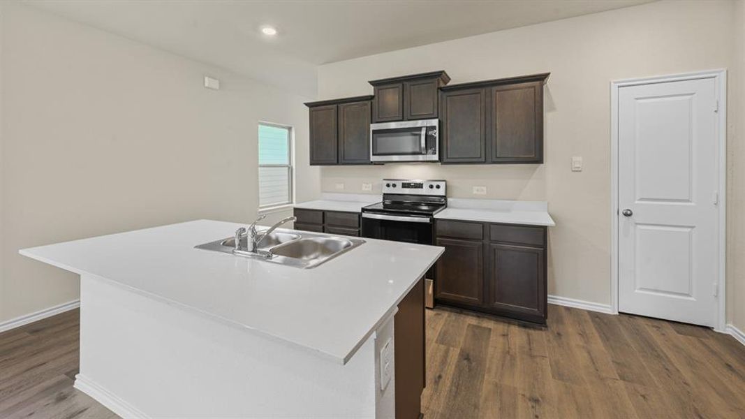 Kitchen featuring a center island with an undermount sink, dark wood cabinetry, white countertops, stainless steel microwave, and wood-finish flooring