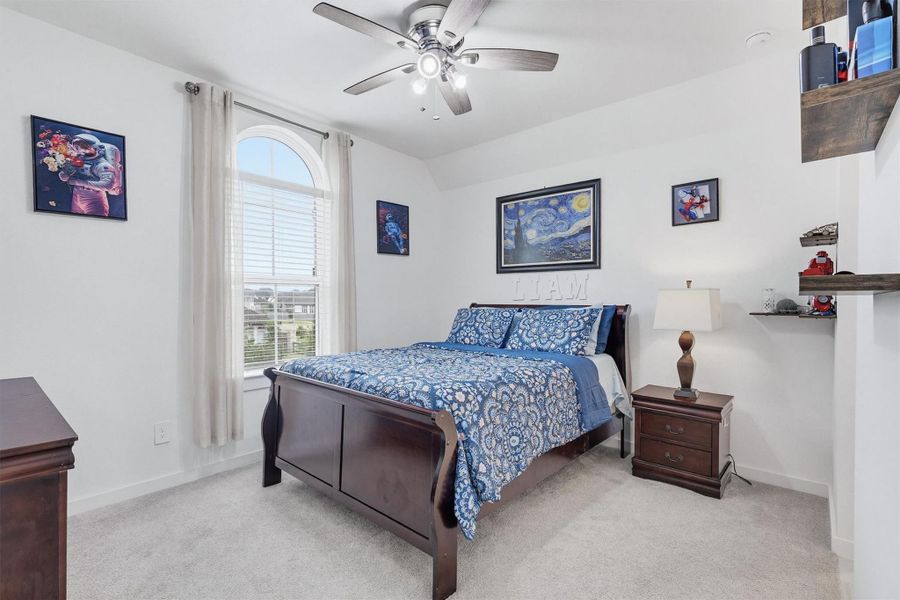 This bedroom features soft carpeting, a ceiling fan, and a window with privacy blinds, creating a comfortable and well-lit space.