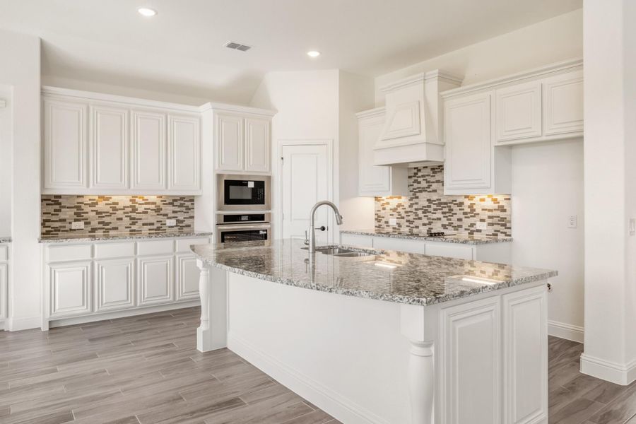 Kitchen with white cabinets, granite island, tile backsplash, and stainless steel appliances on wood-look floors Kitchen with white cabinets, granite island, tile backsplash, and stainless steel appliances on wood-look floors