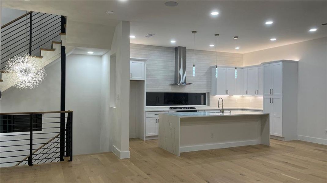 Kitchen with hanging light fixtures, a kitchen island with sink, wall chimney exhaust hood, white cabinetry, and light wood-type flooring