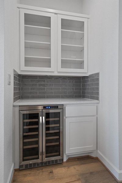 Built-in beverage station featuring a dual-zone wine refrigerator, white shaker-style cabinetry with glass-front uppers, a light-toned countertop, and gray subway tile backsplash