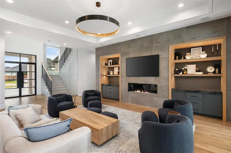 Sophisticated living room featuring a striking floor-to-ceiling tile fireplace, custom built-in shelving with accent lighting, and a sleek linear gas fireplace. Real hardwood floors and a modern statement chandelier complete the space, creating a warm yet contemporary setting perfect for relaxing or entertaining.