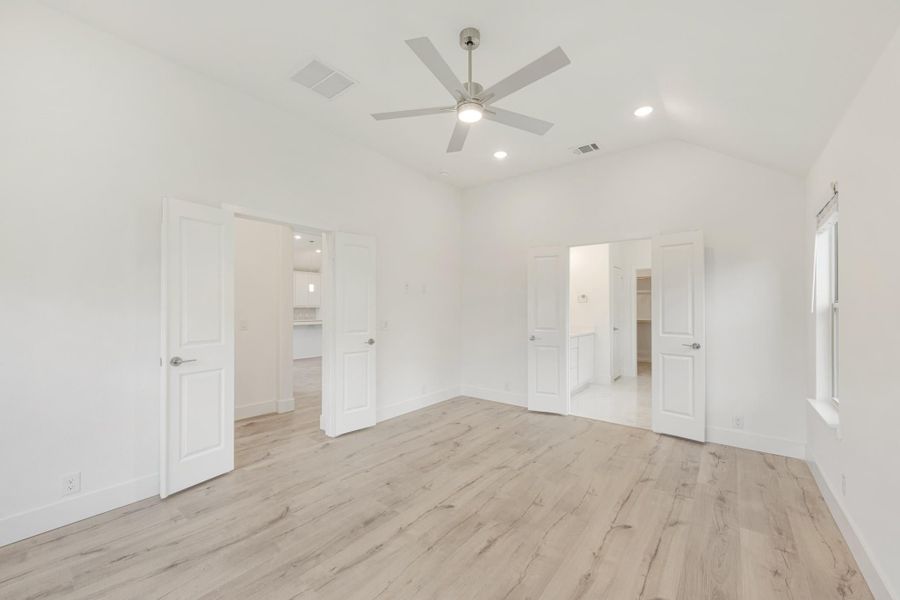 Empty bedroom with vaulted ceiling, ceiling fan, light wood floors, and doors leading to bathroom and closet.