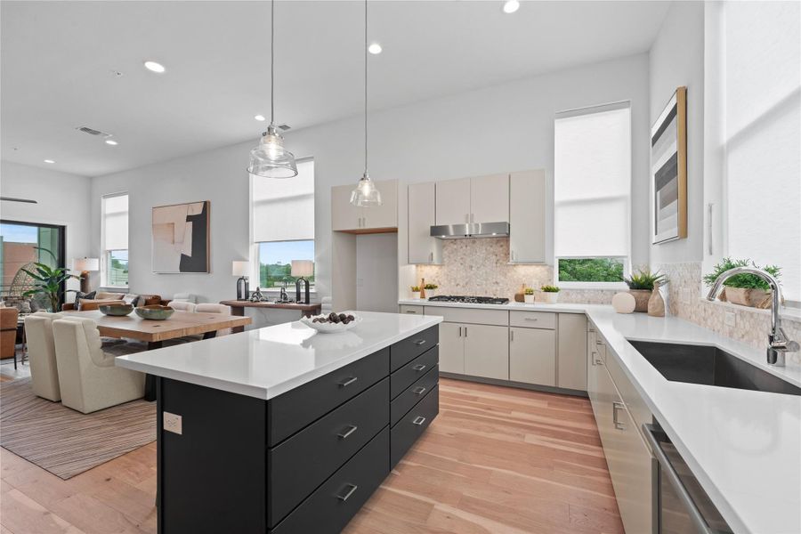 Open-concept kitchen featuring a large island with white countertop, light wood-finish flooring, white cabinetry, a tile backsplash, and a stainless steel sink with a high-arc faucet