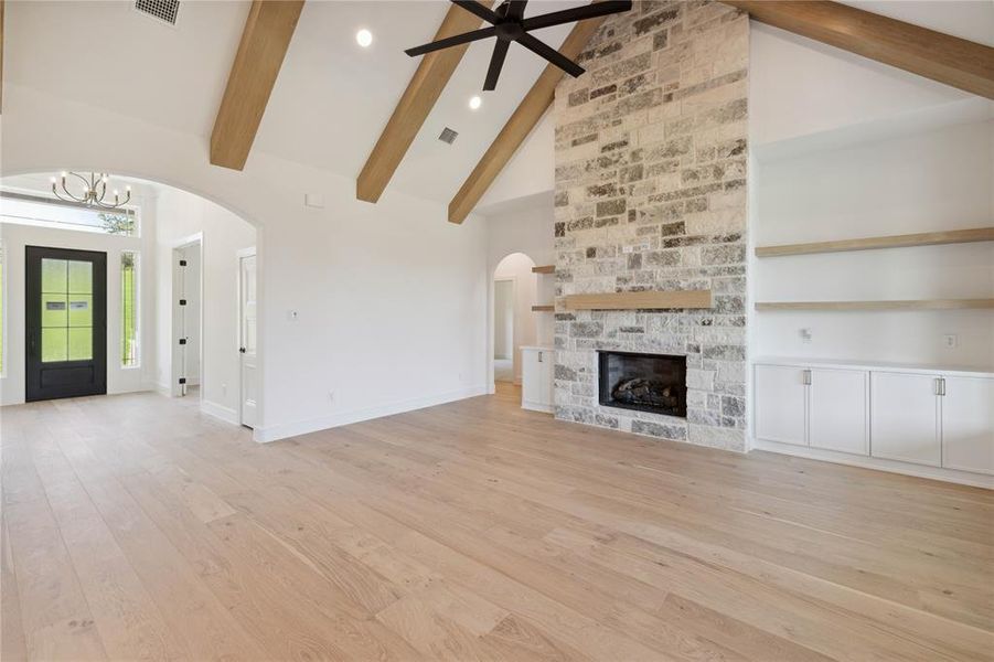 Unfurnished living room featuring a stone fireplace, ceiling fan with notable chandelier, high vaulted ceiling, beam ceiling, and light wood-type flooring