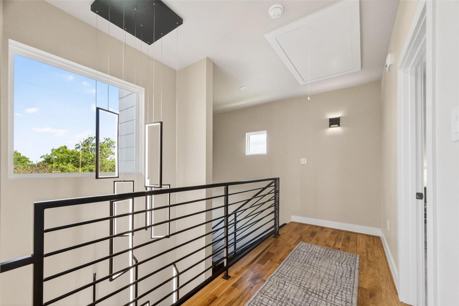 The third-floor landing is a well-lit and thoughtfully finished transitional space, with hardwood floors, a large window, black metal railings, and a designer pendant fixture. It connects the primary suite, secondary bedroom, and utility room with ease.